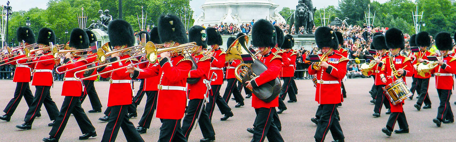 Marching band of British soldiers in their iconic red uniforms marching around the streets near Buckingham Palace. There are crowds gathered on the sides.
